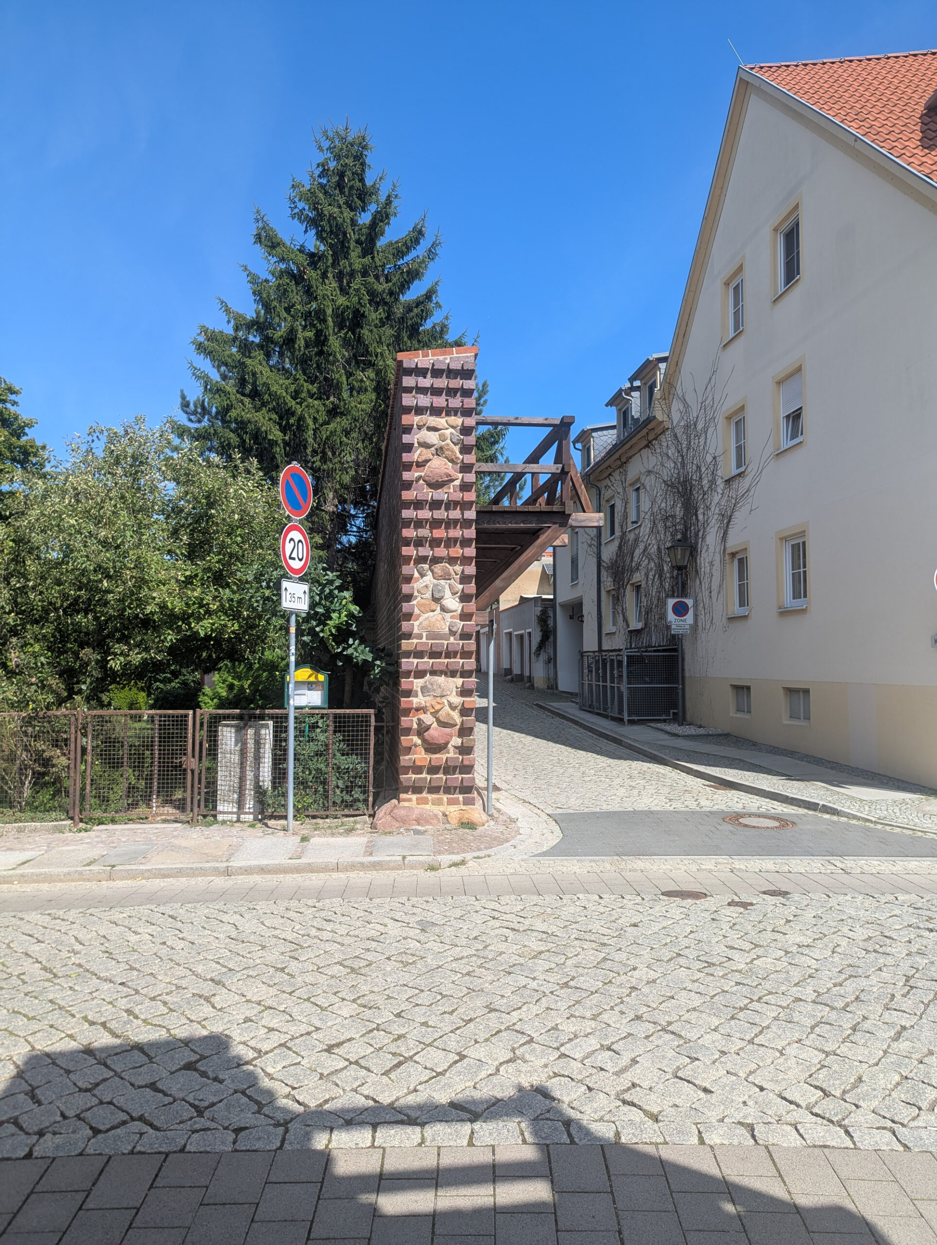 Ein rotes Tempolimit-Schild (20 km/h) steht an einer Straßenecke in Delitzsch, Sachsen. Die Straße führt leicht bergauf und ist von Wohnhäusern gesäumt. Das Bild zeigt einen sonnigen Tag mit deutlichen Schatten.