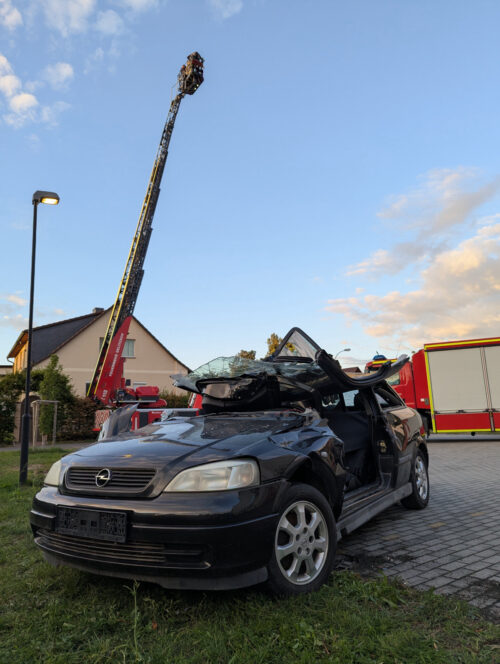 Ein stark beschädigter Pkw steht beim Feuerwehr-Vereinsfest vor dem Gerätehaus Benndorf; dahinter ist die ausgefahrene Drehleiter zu sehen.
