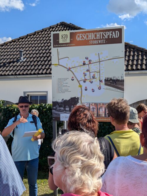 Ein Mann mit Hut erklärt einer Besuchergruppe die Übersichtstafel mit den Stationen des Storkwitzer Geschichtspfads.